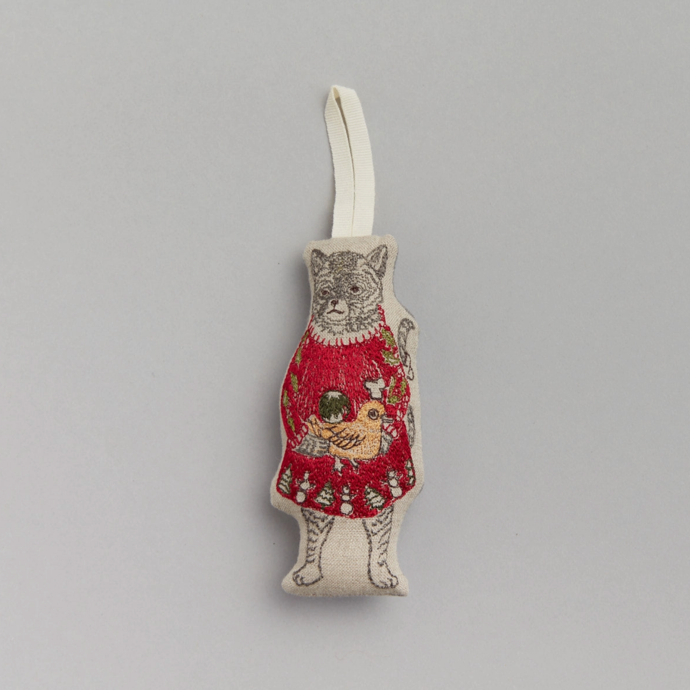 A fabric ornament depicting a embroidered cat in a red Christmas dress holding presents, with a white hanging loop attached.