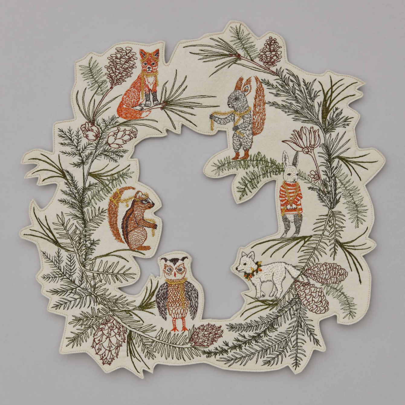 A round felt wreath with embroidered pine needles and Christmas animal characters including a fox, squirrel, bunny, arctic fox, owl, and chipmunk, dressed in metallic gold tinsel, among pine cones and branches.