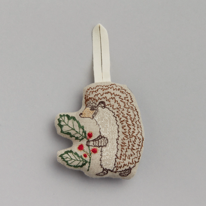 A fabric hedgehog ornament with a brown textured coat, holding a branch with holly leaves and red berries, designed for holiday decor.