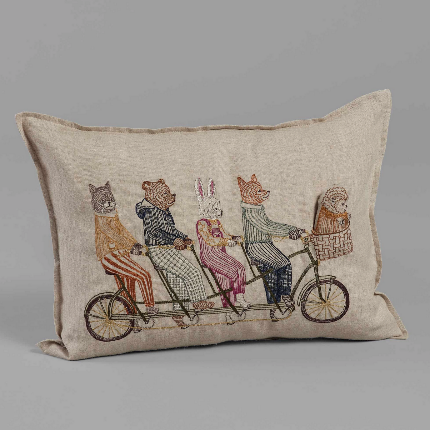 A decorative pillow with an embroidered of a fox, bunny, bear, and cat on a tandem bike, with a hedgehog in the basket.