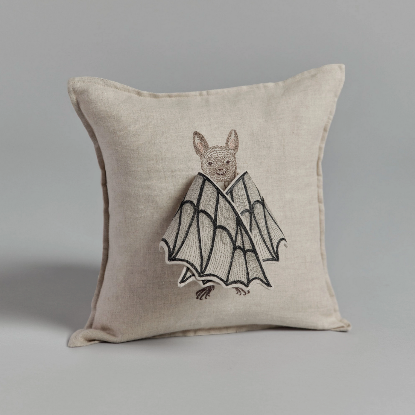 A square beige pillow with a bat design that has its wings spread open, revealing a butterfly behind.
