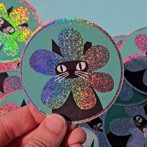 A round sticker with a design of a black cat wearing a multicolored sparkly flower around its face. The sticker is held between fingers against a teal background.