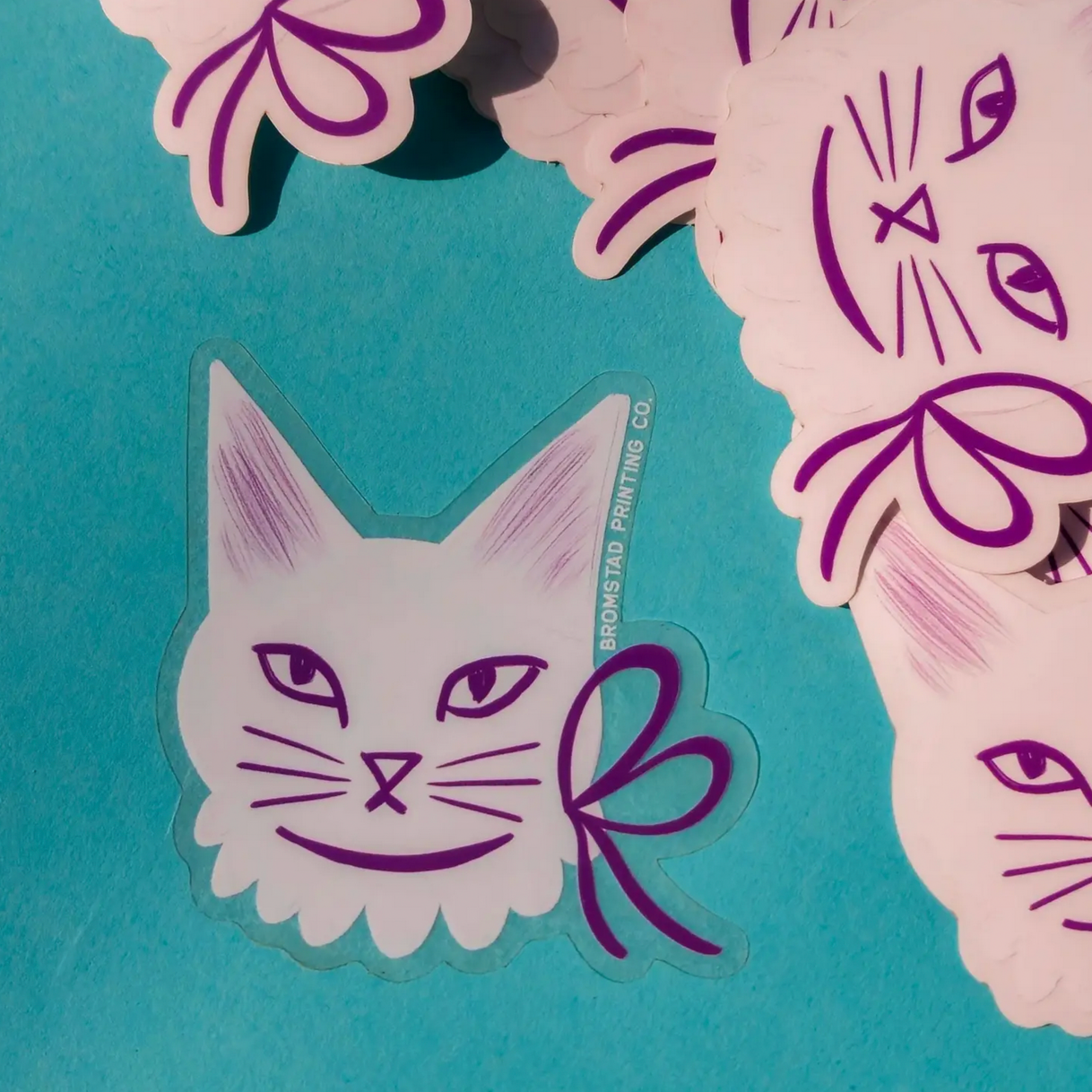 Clear stickers with a design of a white cat wearing a purple ribbon around its neck, displayed on a blue background.