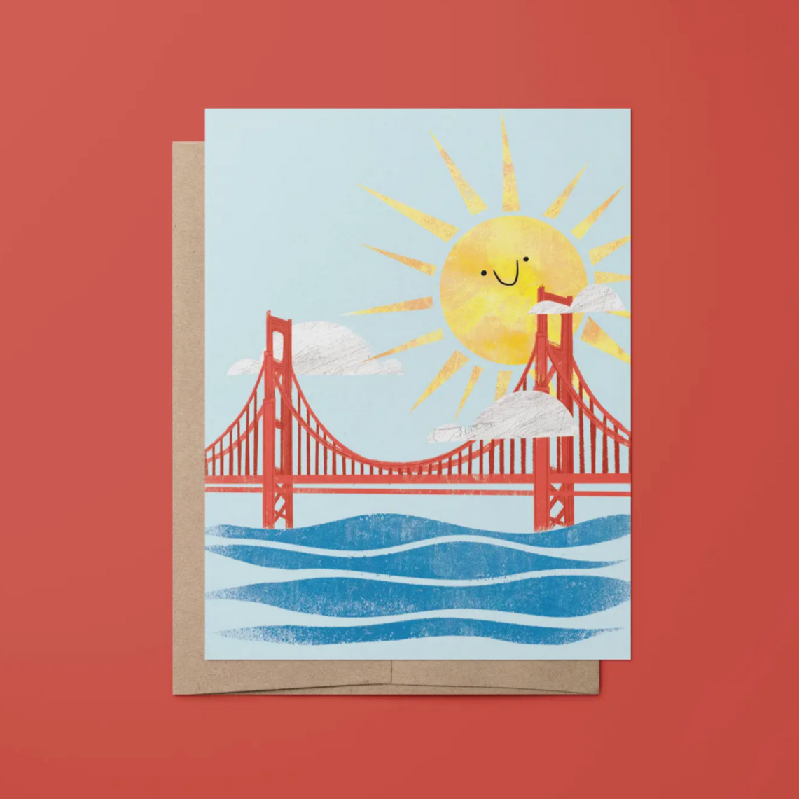 A greeting card with an illustration of the Golden Gate Bridge in San Francisco, featuring a smiling sun and blue sky. The card is shown with a kraft paper envelope against a red background.