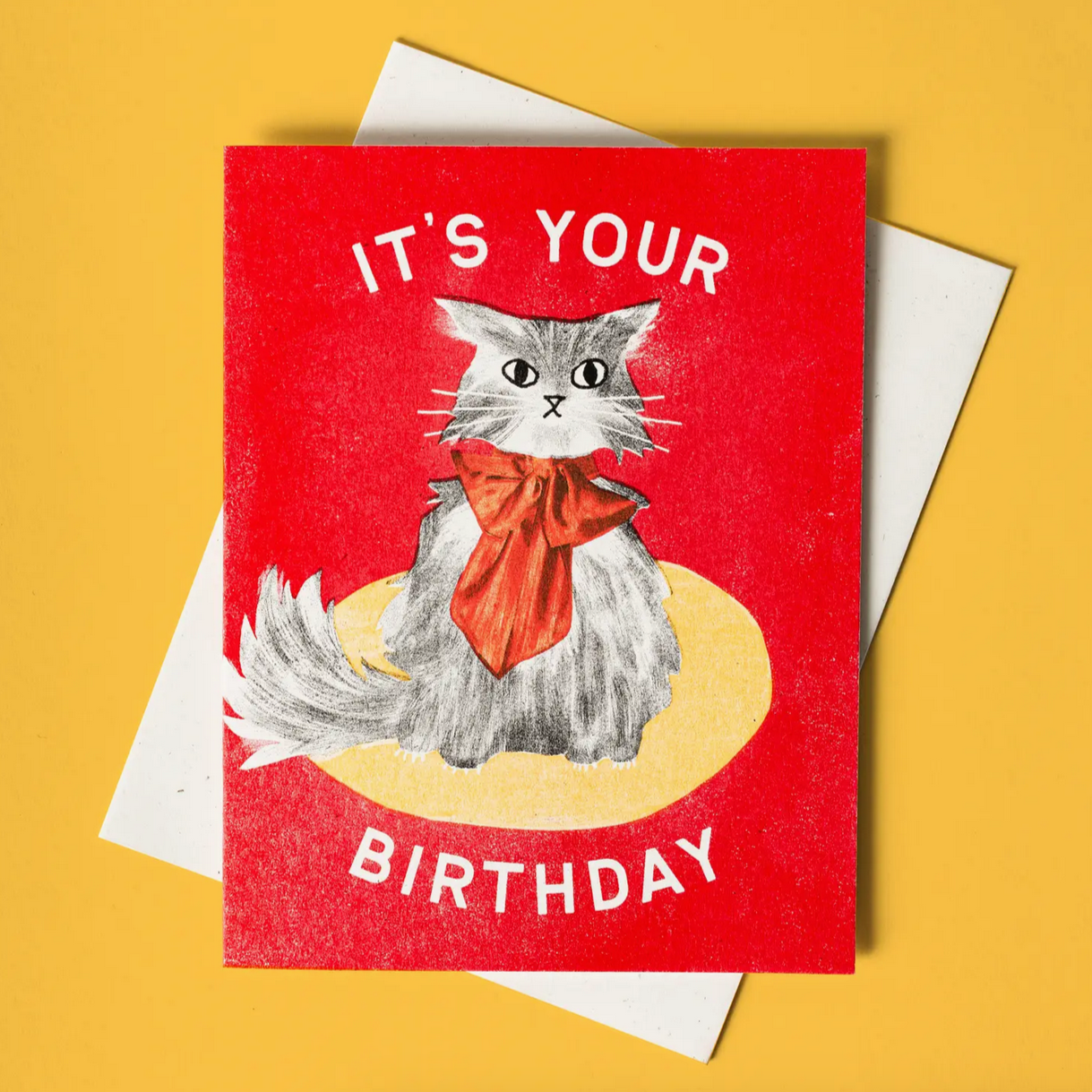 A birthday card with an illustration of a cat wearing a bow tie, with the text 'IT'S YOUR BIRTHDAY' on the front. There is a white envelope included, placed behind the card.