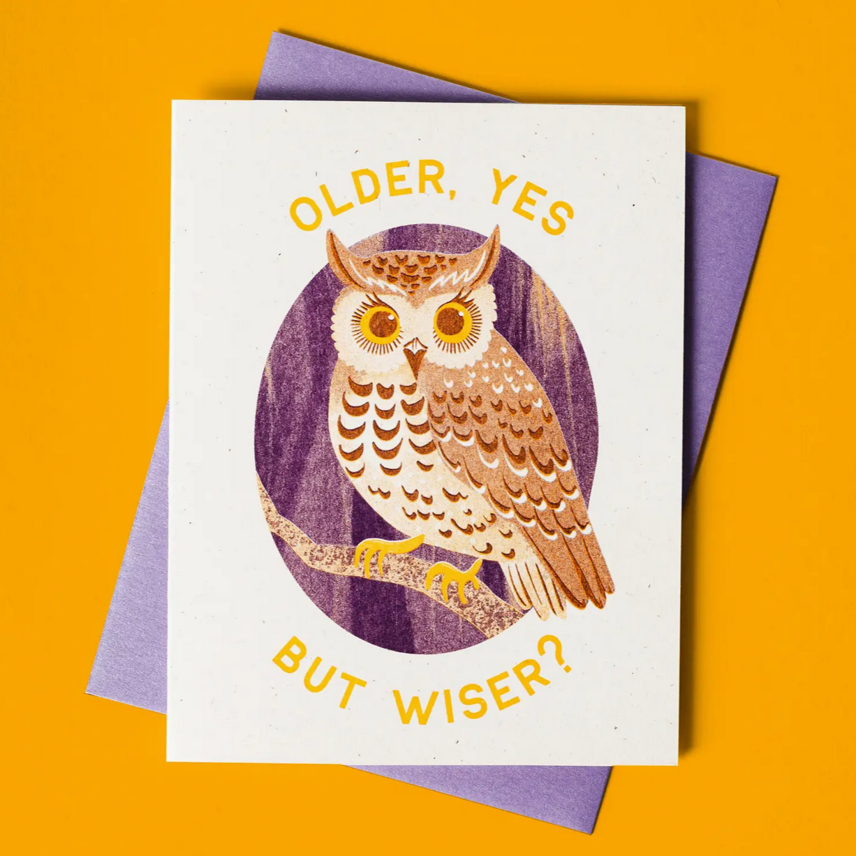 A greeting card with an illustration of an owl and the text 'OLDER, YES BUT WISER?' printed on it. The card is displayed with a purple envelope on a yellow background.
