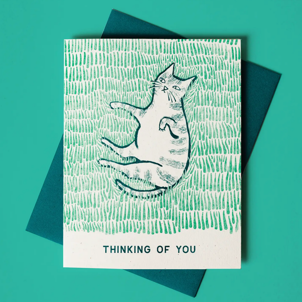 A Risograph printed greeting card depicting a stylized cat lying down with the text 'THINKING OF YOU' at the bottom, accompanied by a teal envelope.
