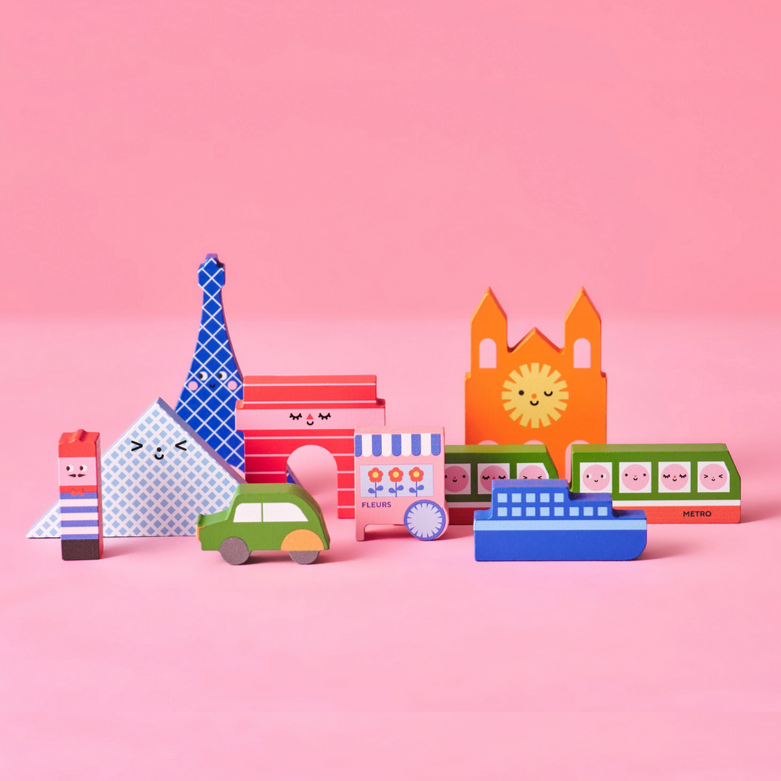 A set of colorful mini wooden city blocks, including iconic landmarks such as a Eiffel Tower and a sun-shaped building, displayed against a pink background.