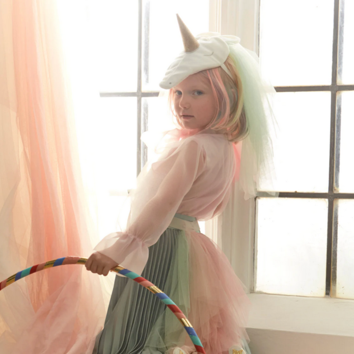 A child wearing a rainbow-colored unicorn costume with a headpiece and a tail, standing by a window.