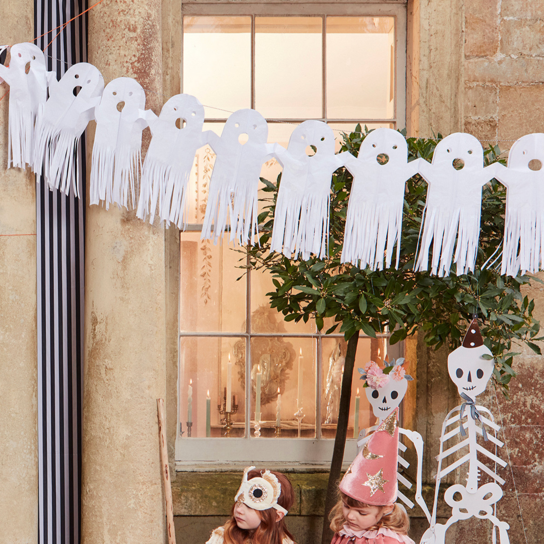 A tissue paper garland of hanging ghosts displayed in front of a window, with children standing and playing with it.