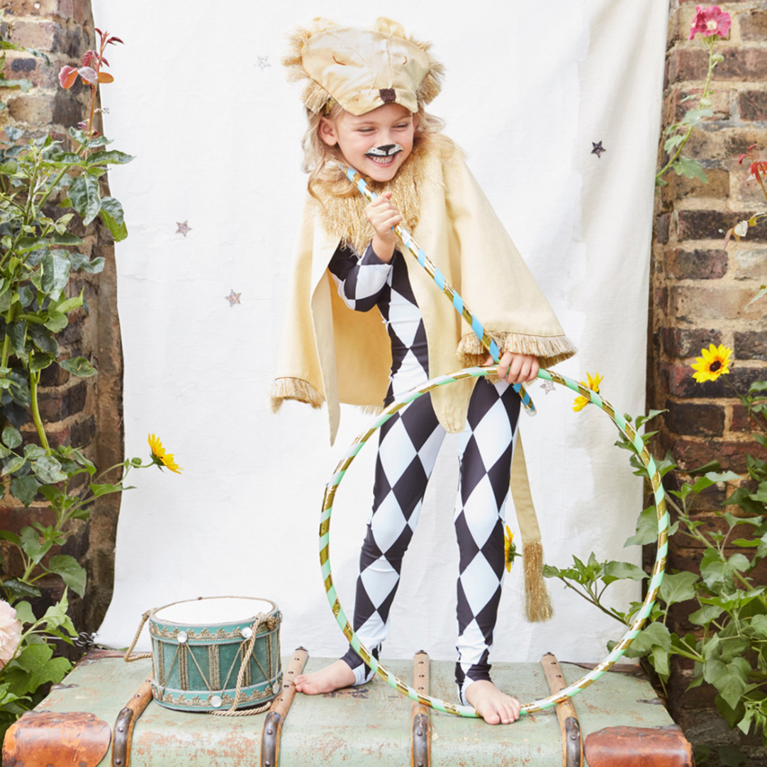 A child wearing a lion costume with a cape and headdress, standing next to a decorative drum set, in a garden setting.