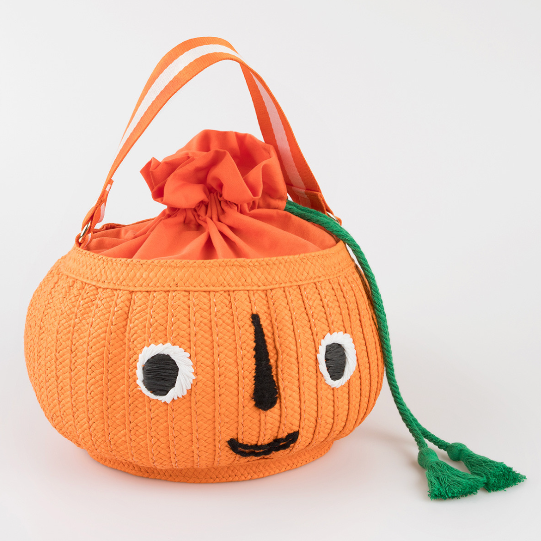 An orange woven paper basket bag with a face detail, green tassels, and an orange drawstring closure. It has two handles and is designed for Halloween.