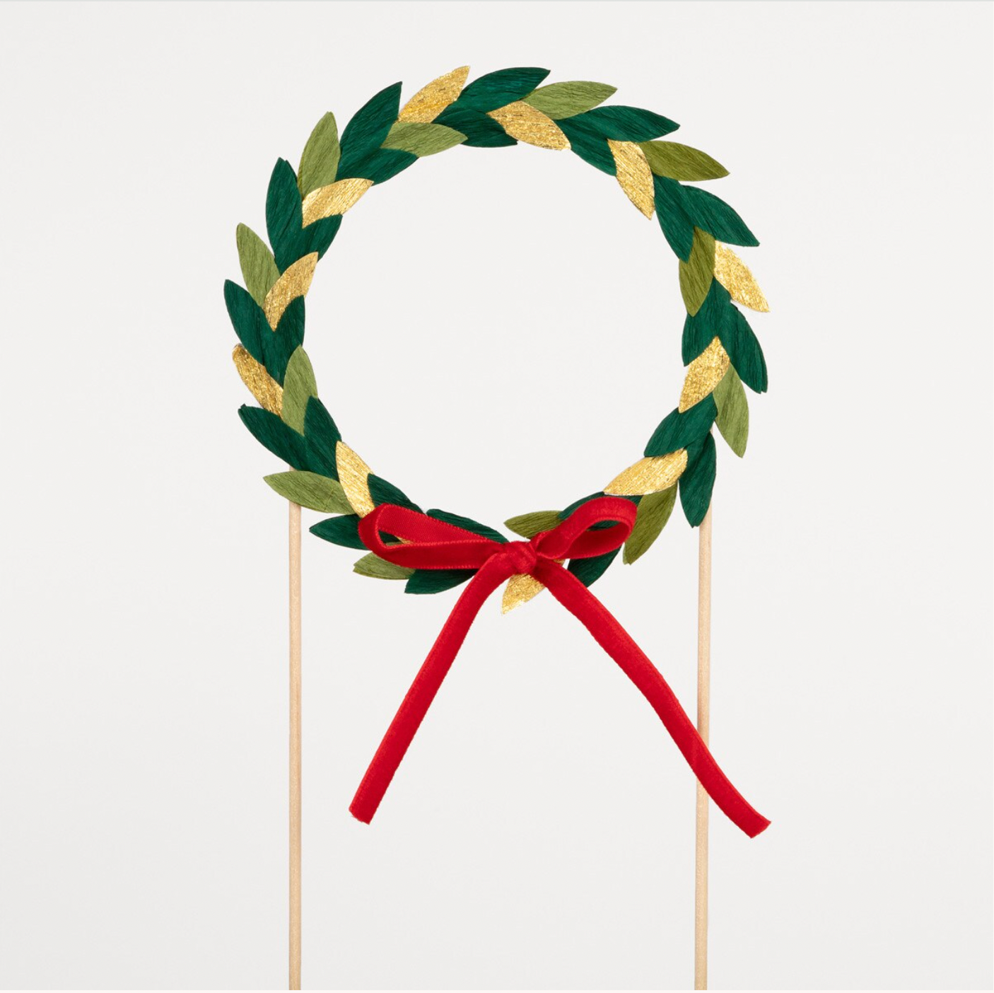 A green, gold, and red crepe paper leaf wreath with a red velvet ribbon, mounted on natural bamboo skewers, designed as a cake topper.