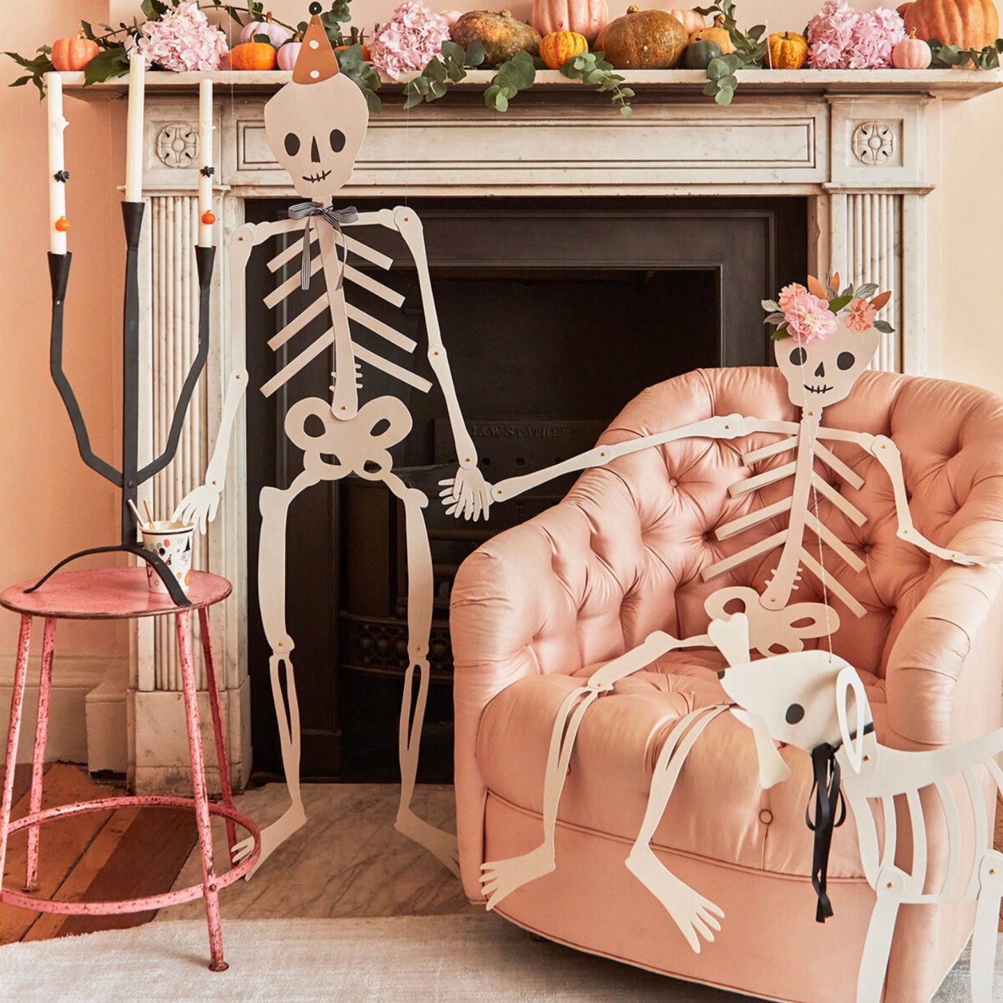 A set of three jointed skeleton decorations displayed in a room, with one skeleton wearing a party hat and satin ribbon bow, another with a floral headpiece and tissue paper flower detail, and a dog skeleton with a black ribbon necktie and silver hanging cords.