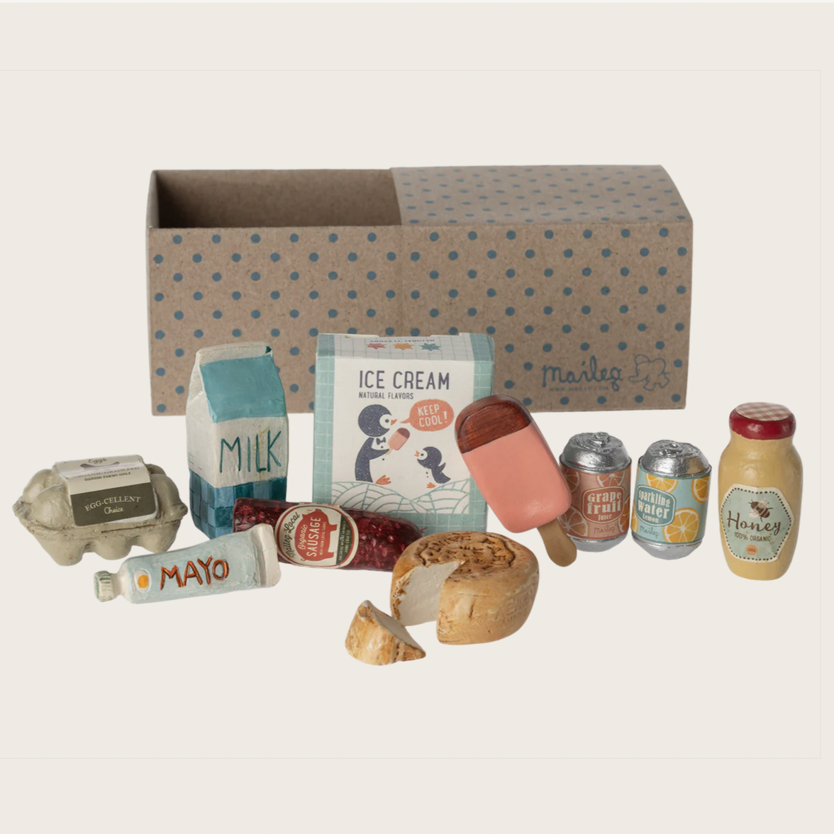 A miniature grocery box with various small toy food items including a carton of milk, a honey jar, sausage, ice cream sticks, egg cartons, soda cans, and cheese, all displayed in front of a cardboard box with 'Maileg' branding.