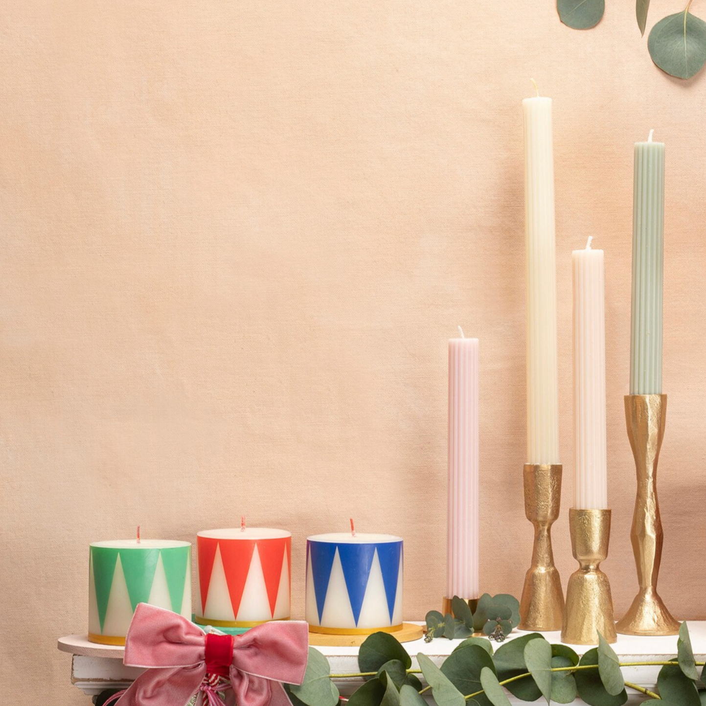 A set of three cylindrical candles with geometric patterns in red, green, and blue colors, featuring gold ink dipped bases and red wicks, displayed on a surface with a pink background and decorative leaves.