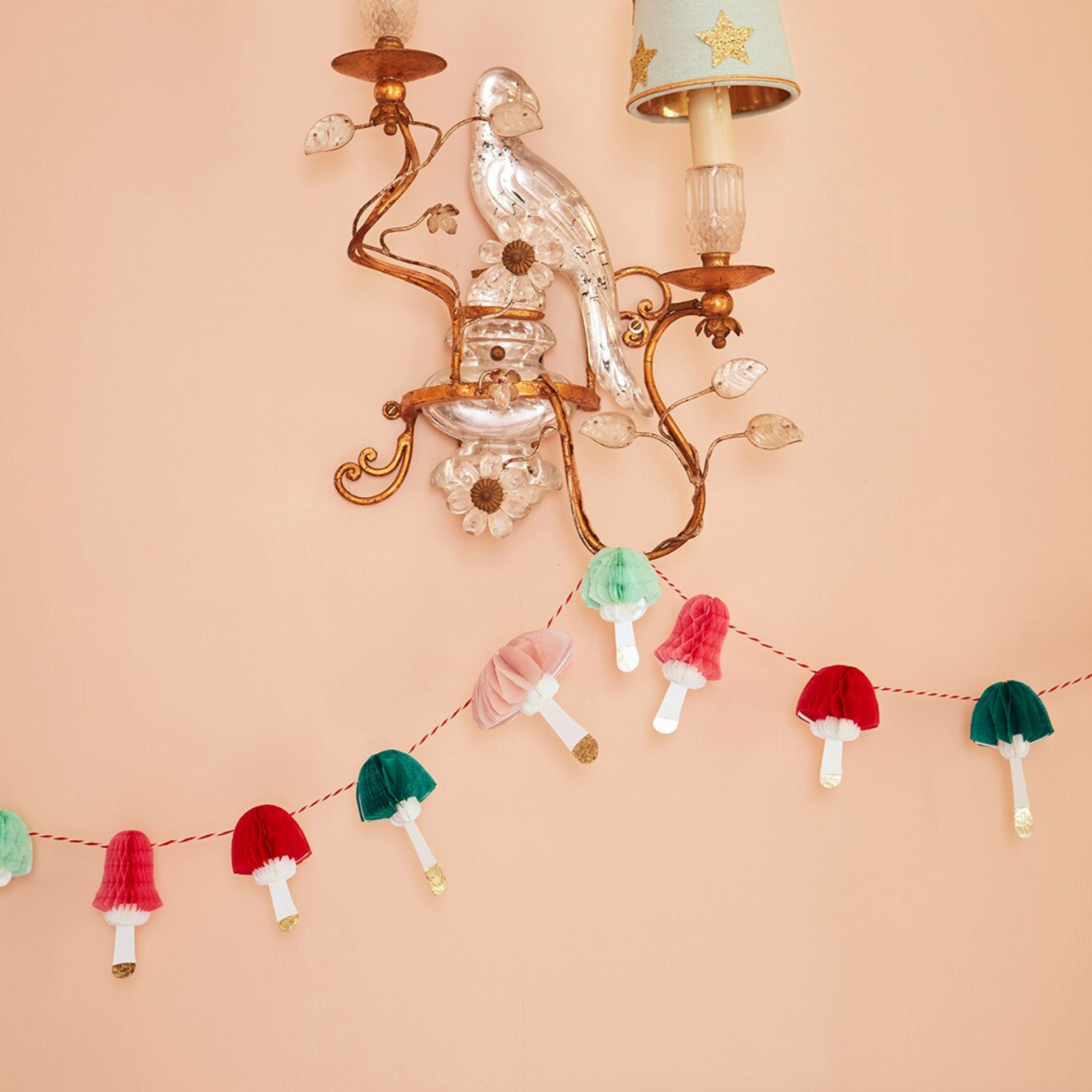 A colorful tissue paper honeycomb mushroom garland with shiny gold foil bases, displayed hanging on a wall sconce.