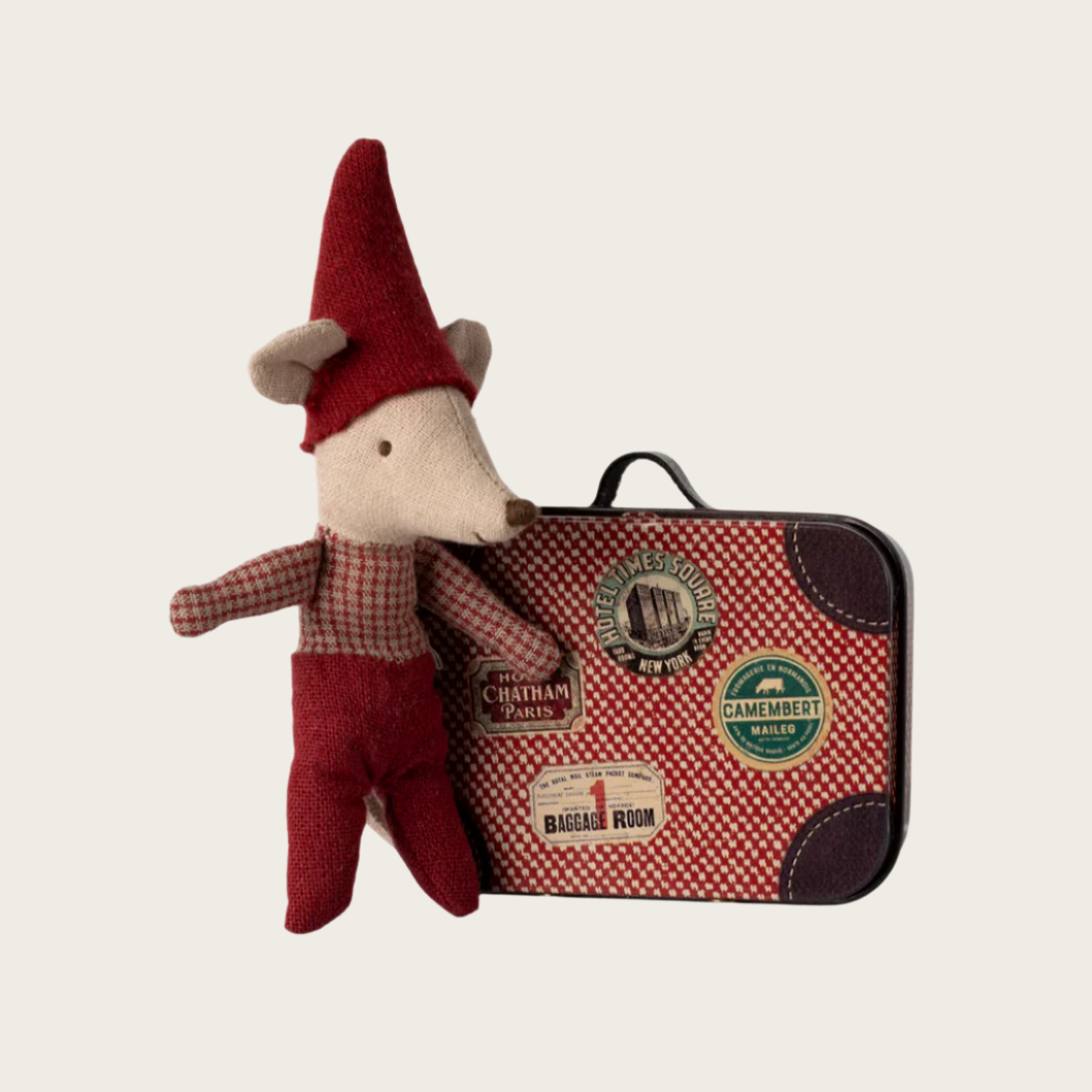 A small toy mouse in a red Christmas outfit sitting next to a red and beige suitcase with travel stickers.
