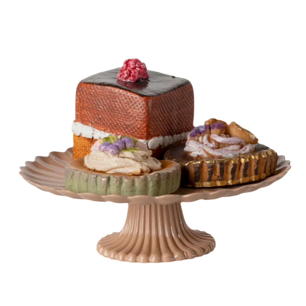 A set of three mini decorated cakes on a rose-colored cake stand, with one cake topped with a red berry, another with a green and purple flower, and the third with a golden, crêpe-like topping.