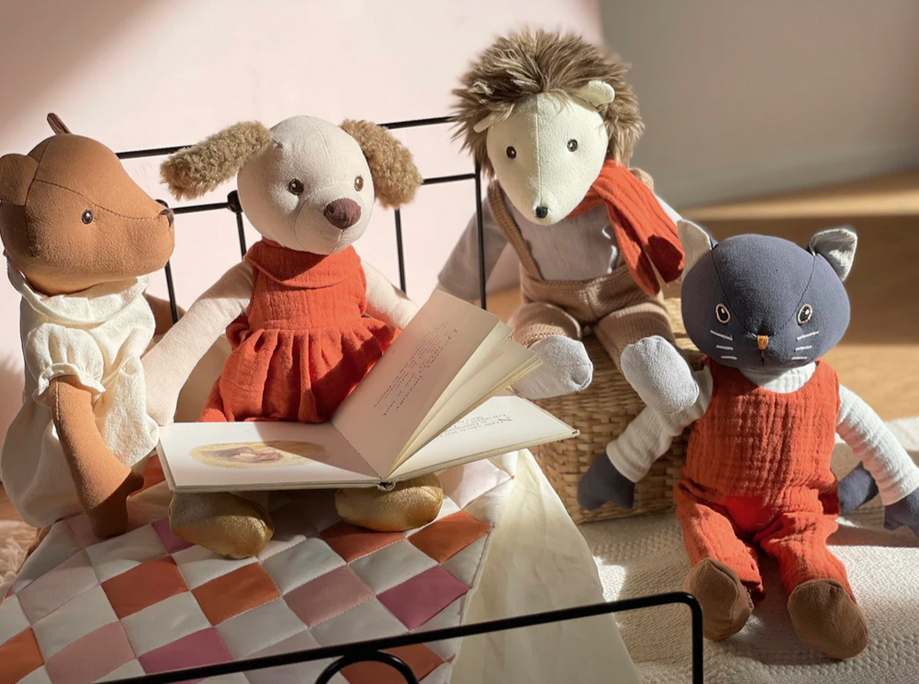 Four plush toys, including a dog, cat, and mouse, sitting on a checkered blanket with an open book.