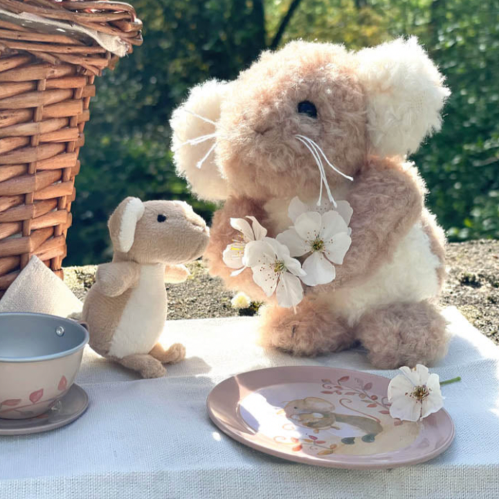 Two stuffed animals, a bear and a mouse, sitting at a table with a picnic setup outdoors.
