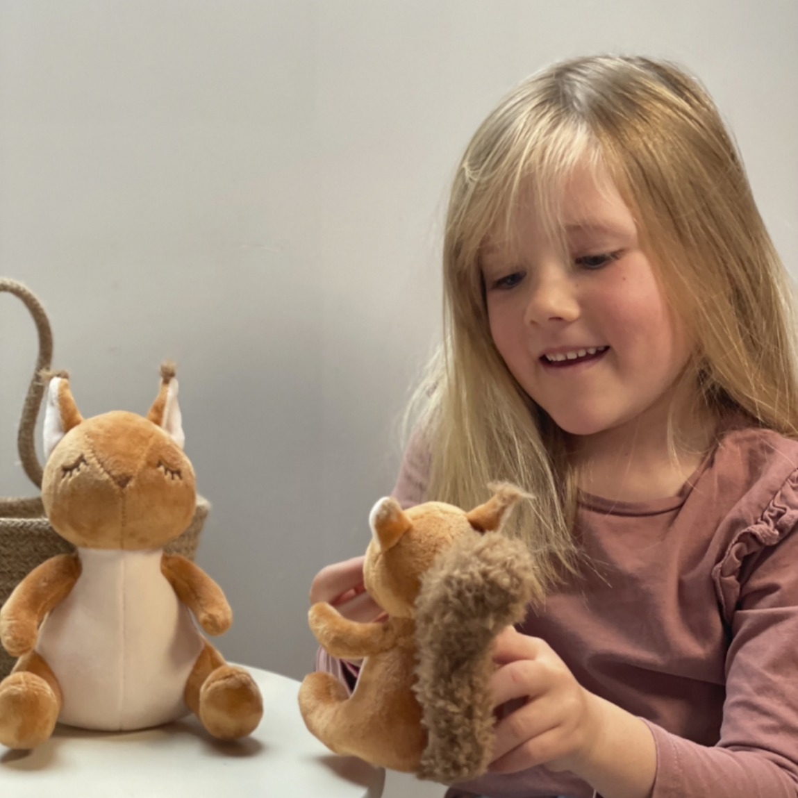 Child holding a plush toy squirrel next to another one on a table.