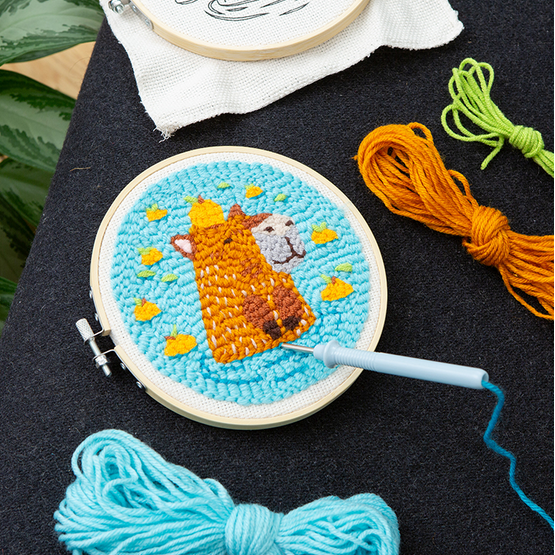 Embroidery hoop with a completed design of a fox and a dog on a blue background, surrounded by yarn and an embroidery hoop.