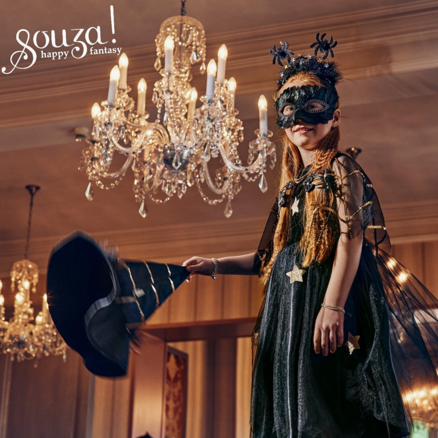 Person in a dark, elaborate costume holding a top hat in an elegant room with chandeliers.