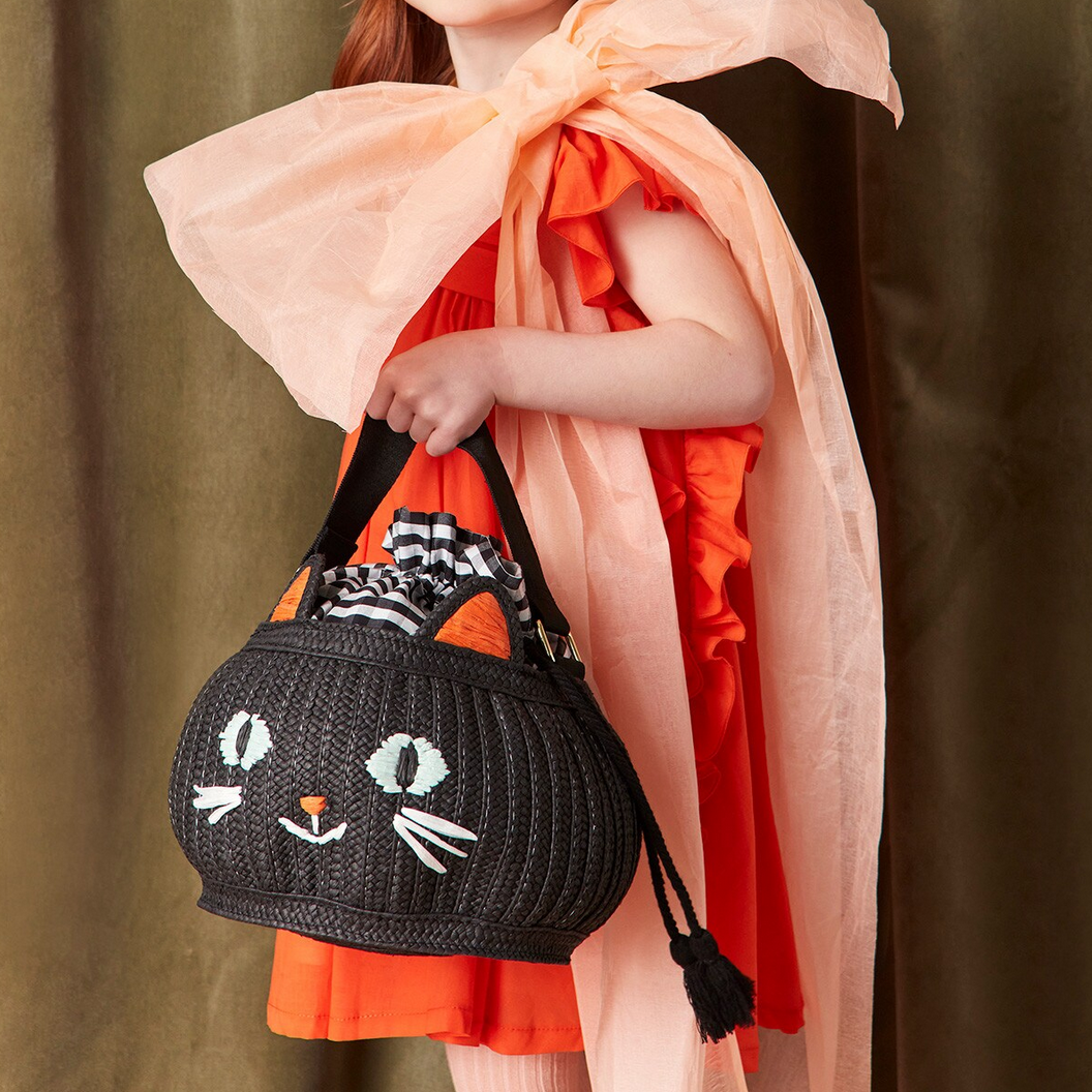 Child holding a black woven handbag with cat face design, wearing an orange dress and pink scarf.