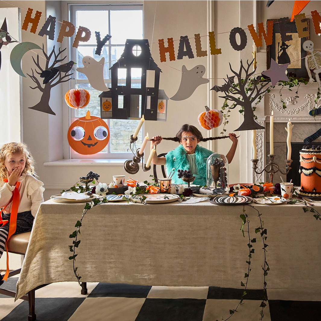 Children at a Halloween-themed table with decorations and 'Happy Halloween' banner.