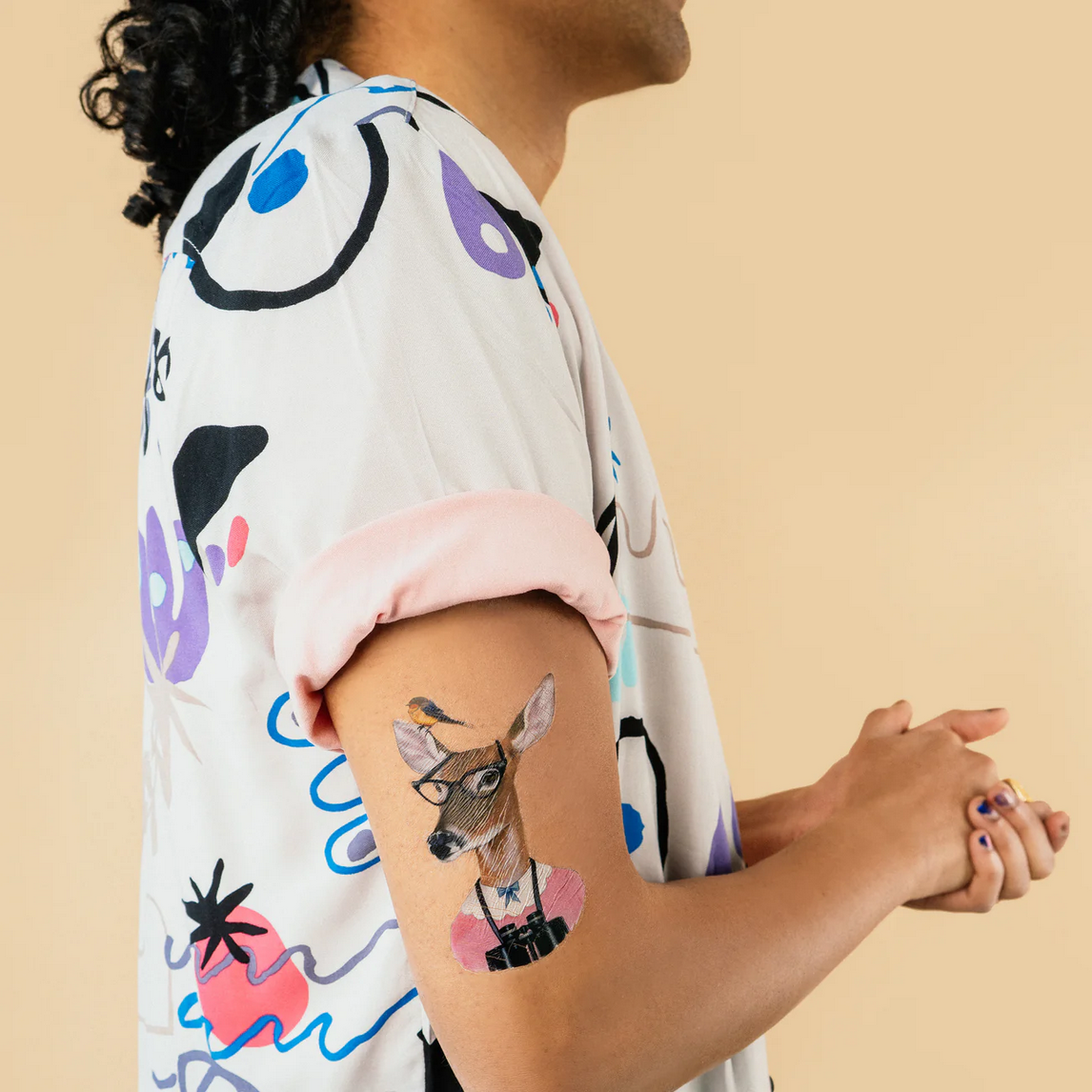 Person wearing a colorful shirt with a deer tattoo on their arm against a beige background