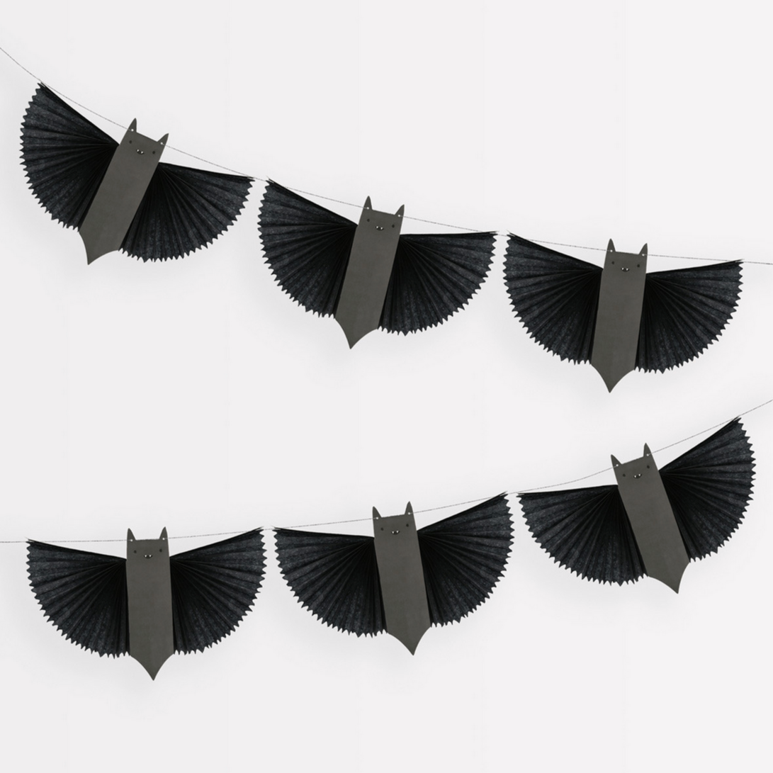 A Halloween bat garland with six black paper bats with concertina wings, strung together with shiny silver metallic cord against a white background.