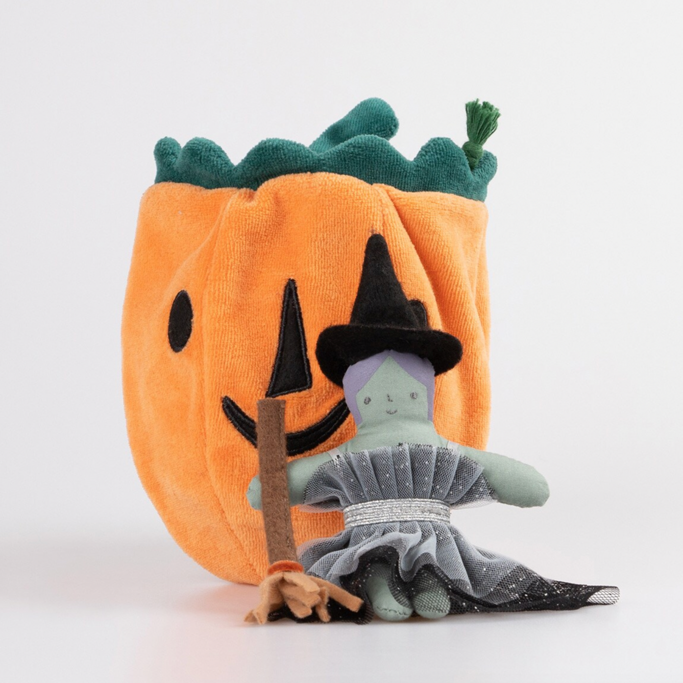 A small doll designed to look like a witch with a green and grey outfit, sitting next to an orange pumpkin-shaped pouch with a face drawstring. The doll has a black and orange hat and a brown and orange broomstick.