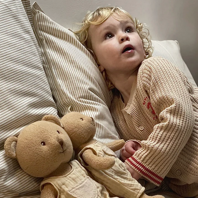 Child lying on a bed with a teddy bear, wearing a beige sweater.