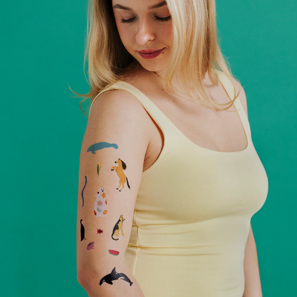 Woman wearing a yellow tank top with various temporary tattoos on her arm against a teal background