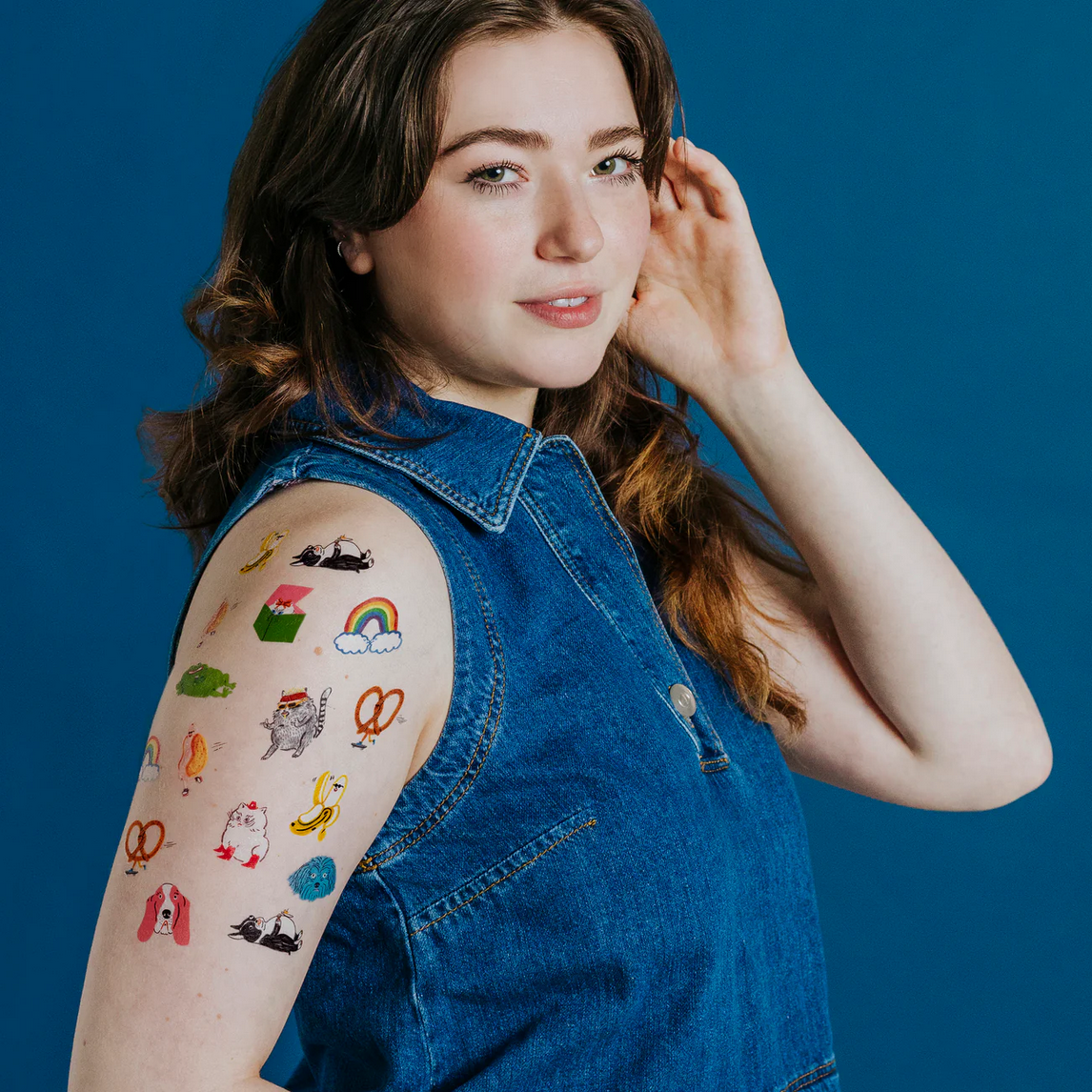 Woman wearing a denim vest with colorful temporary tattoos on her arm against a blue background