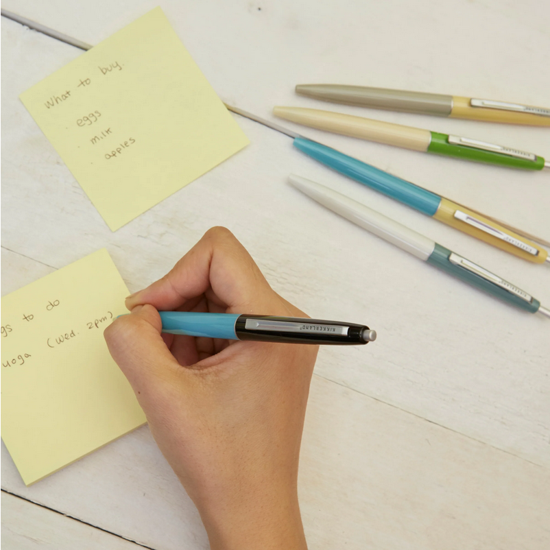 Hand holding a blue pen with sticky notes and other pens on a light surface