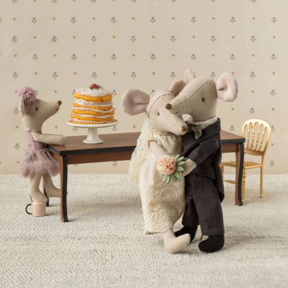 Two toy mice in wedding attire standing in a room with a cake on a table.