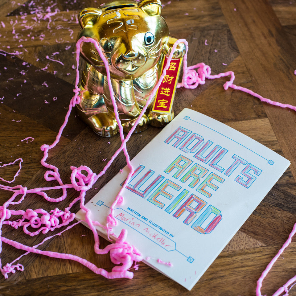 Gold cat with pink string and book titled 'Adults Are Weird' on a wooden surface