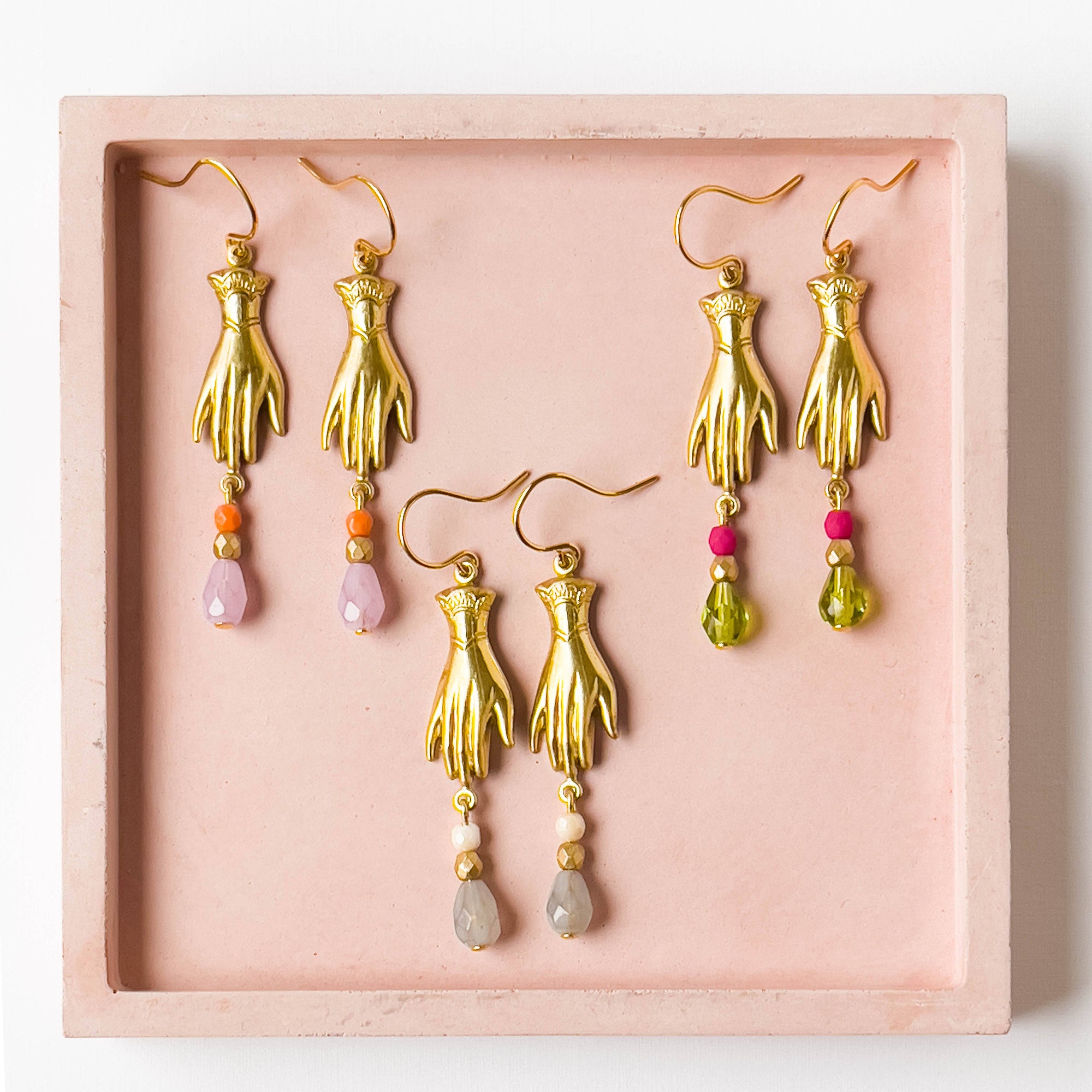 Gold hand-shaped earrings with colorful beads on a pink display box.
