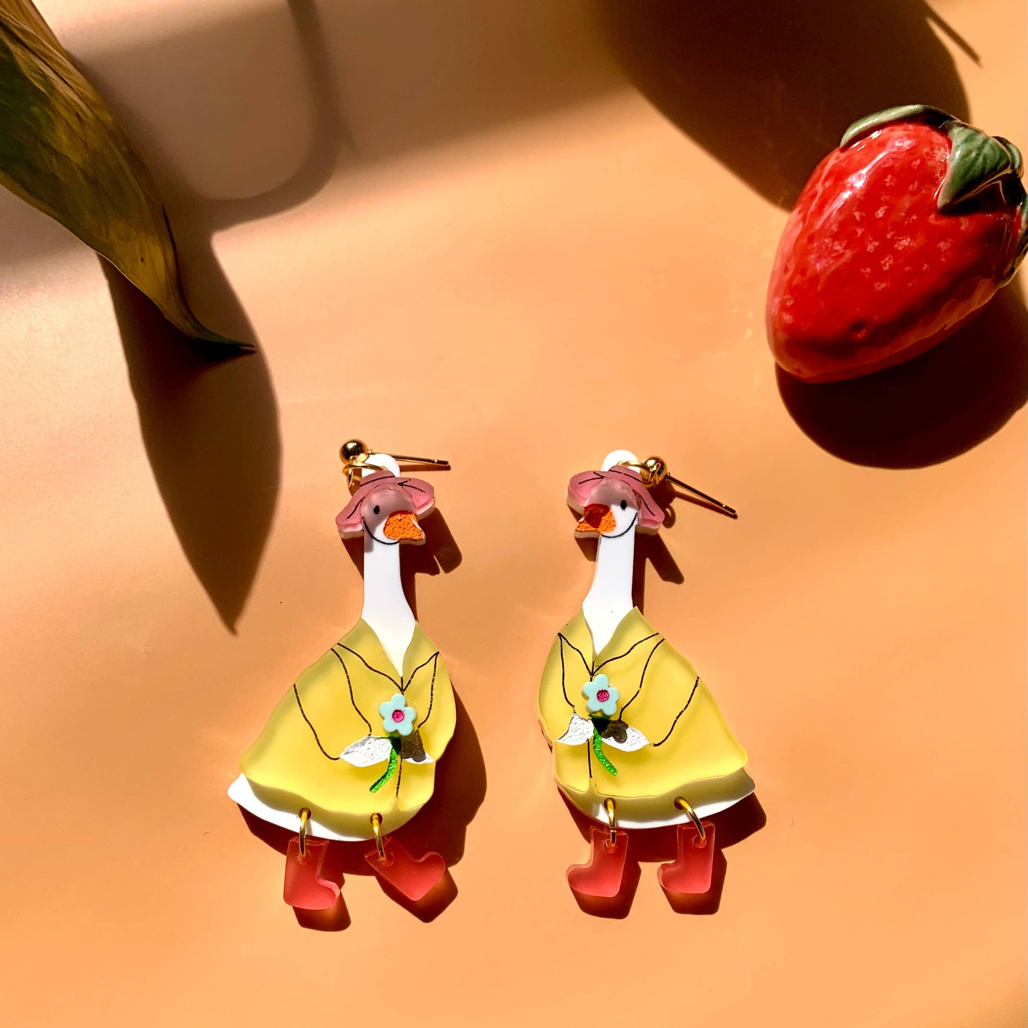 Pair of duck-shaped earrings with yellow dresses on a peach-colored surface.