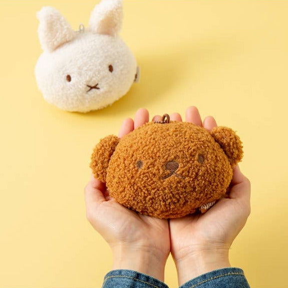 Brown teddy bear-shaped coin purse held by hands with a white rabbit-shaped coin purse in the background on a yellow background.