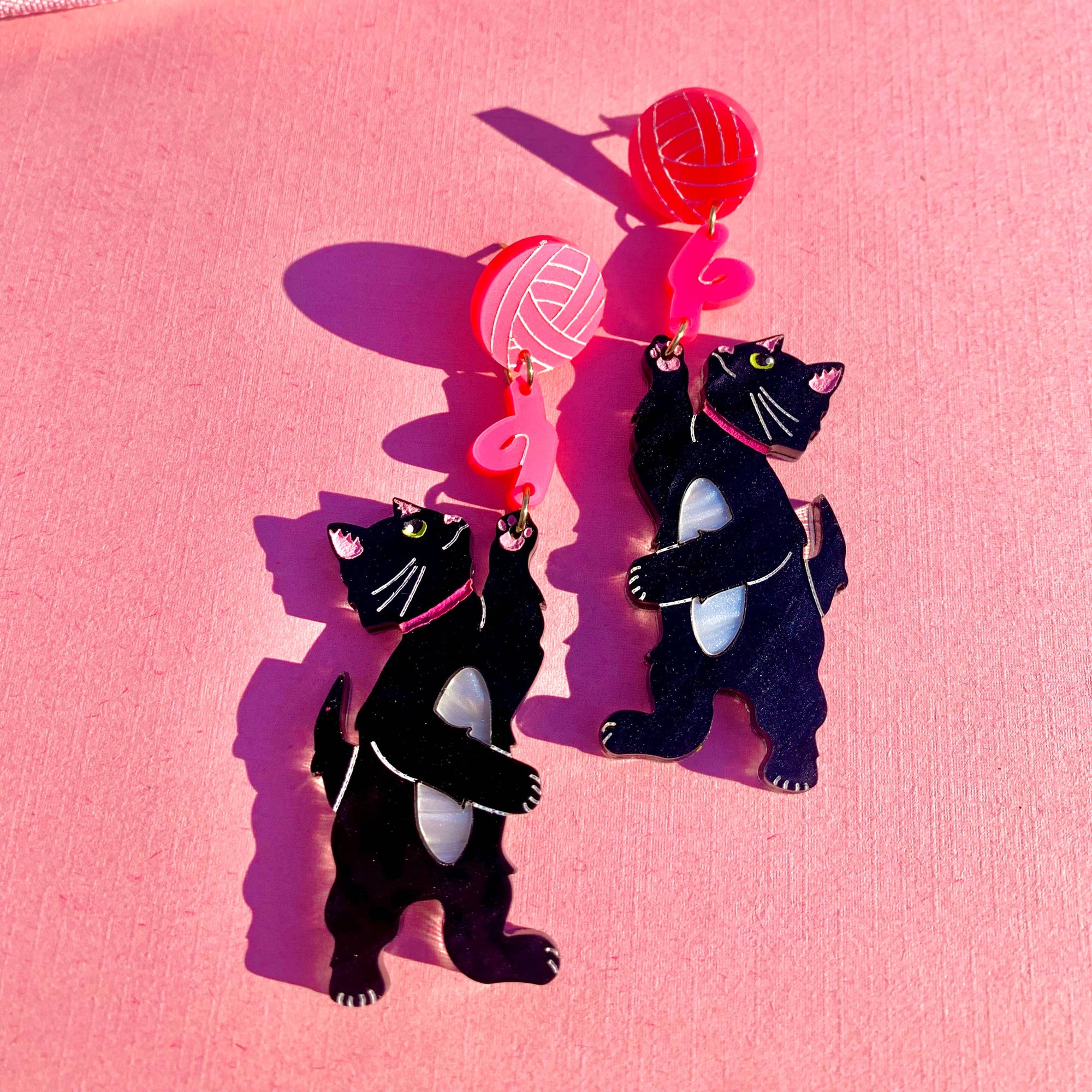 Black cat-shaped earrings with red heart pendants on a pink background