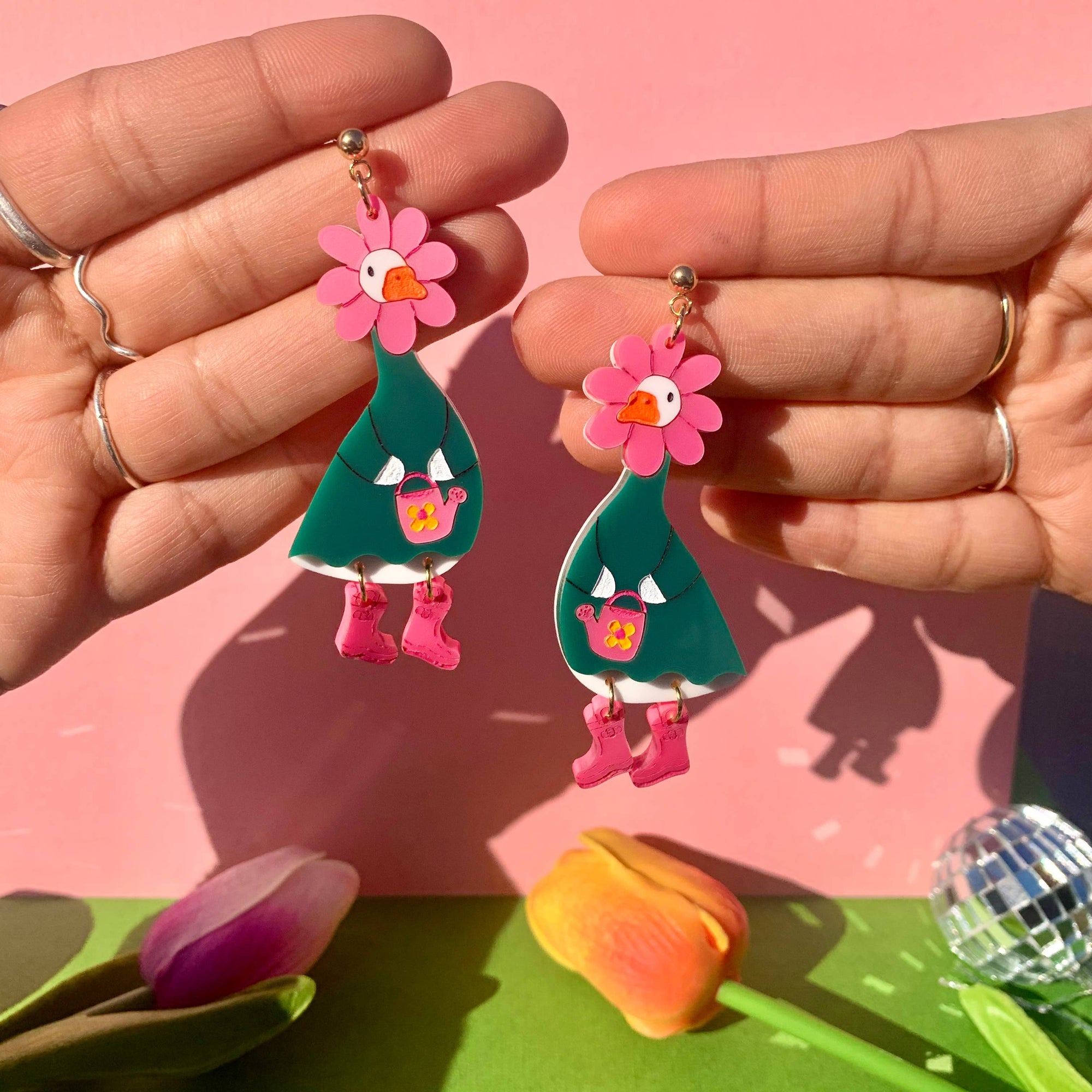 Colorful peacock-shaped earrings held by hands against a pink background with green leaves.