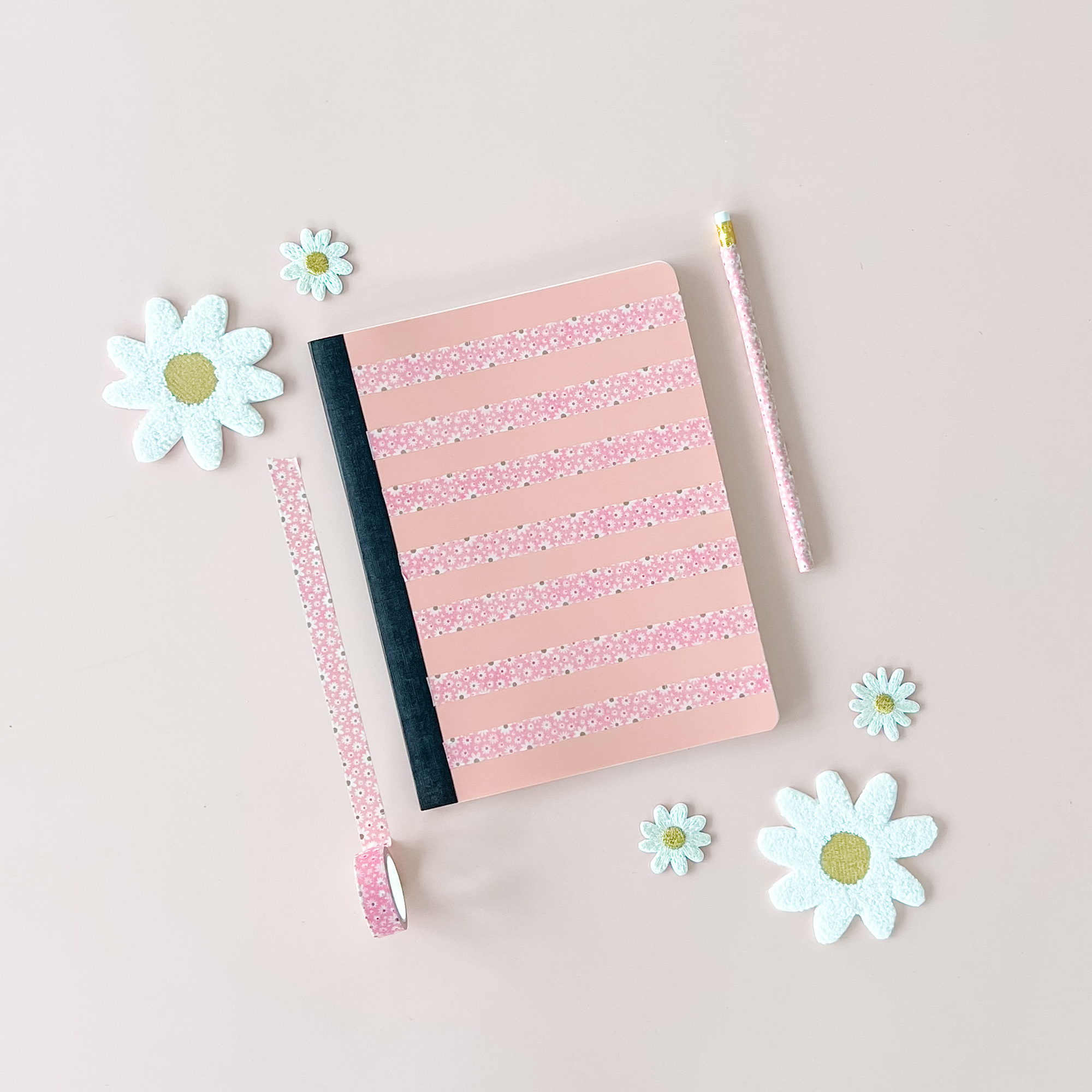 Pink notebook with glittery stripes, a pencil, and decorative flowers on a light beige background