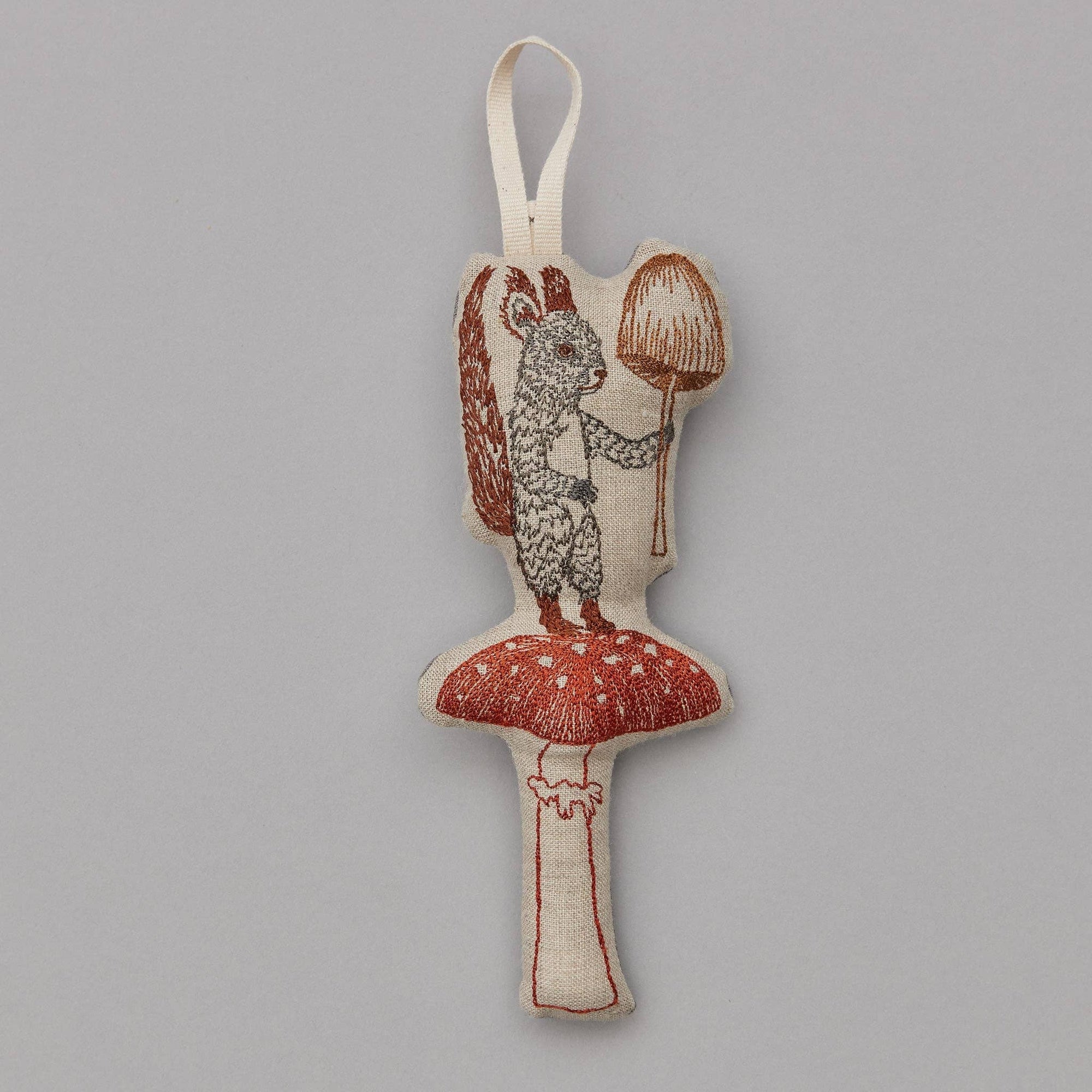 A fabric ornament featuring a gray squirrel holding a red mushroom, attached to a beige loop for hanging.