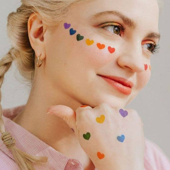Woman with colorful heart tattoos on her face and arm against a neutral background