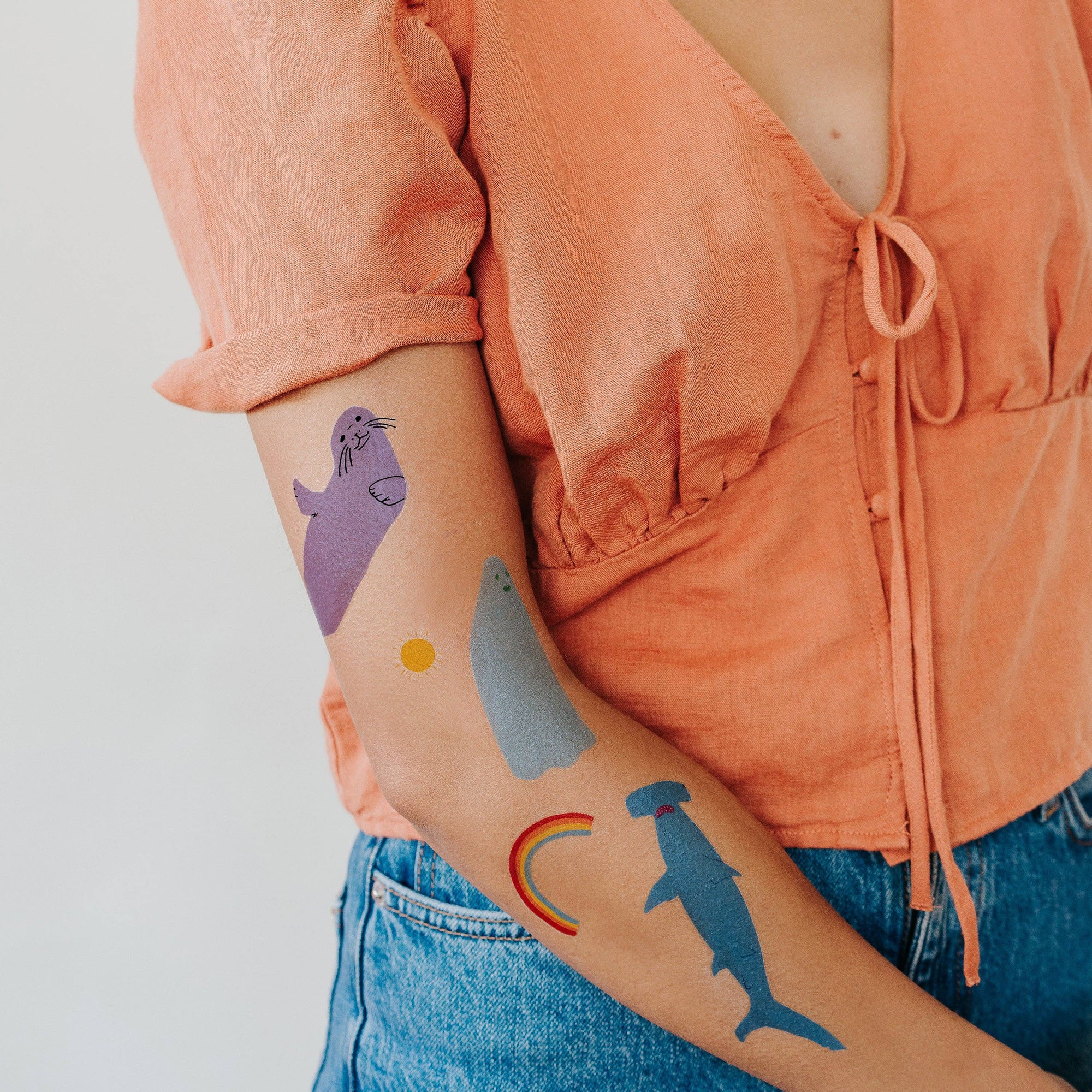 Person wearing temporary tattoos of a purple seal, gray shark, yellow circle, rainbow, and blue dolphin on their arm.