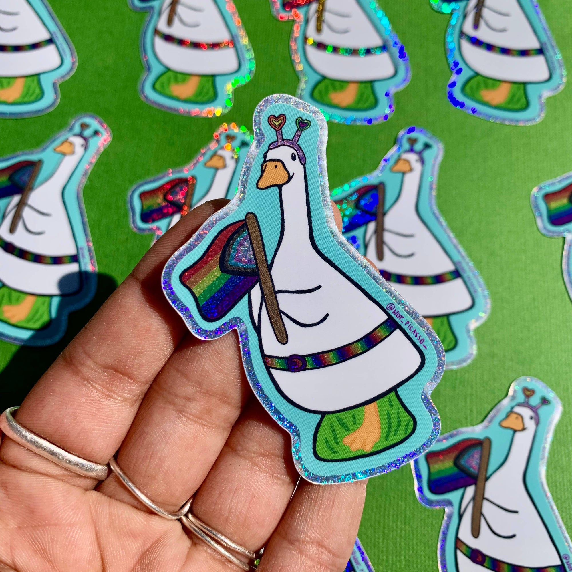 Holographic sticker of a goose holding a rainbow flag on a green background with more stickers.