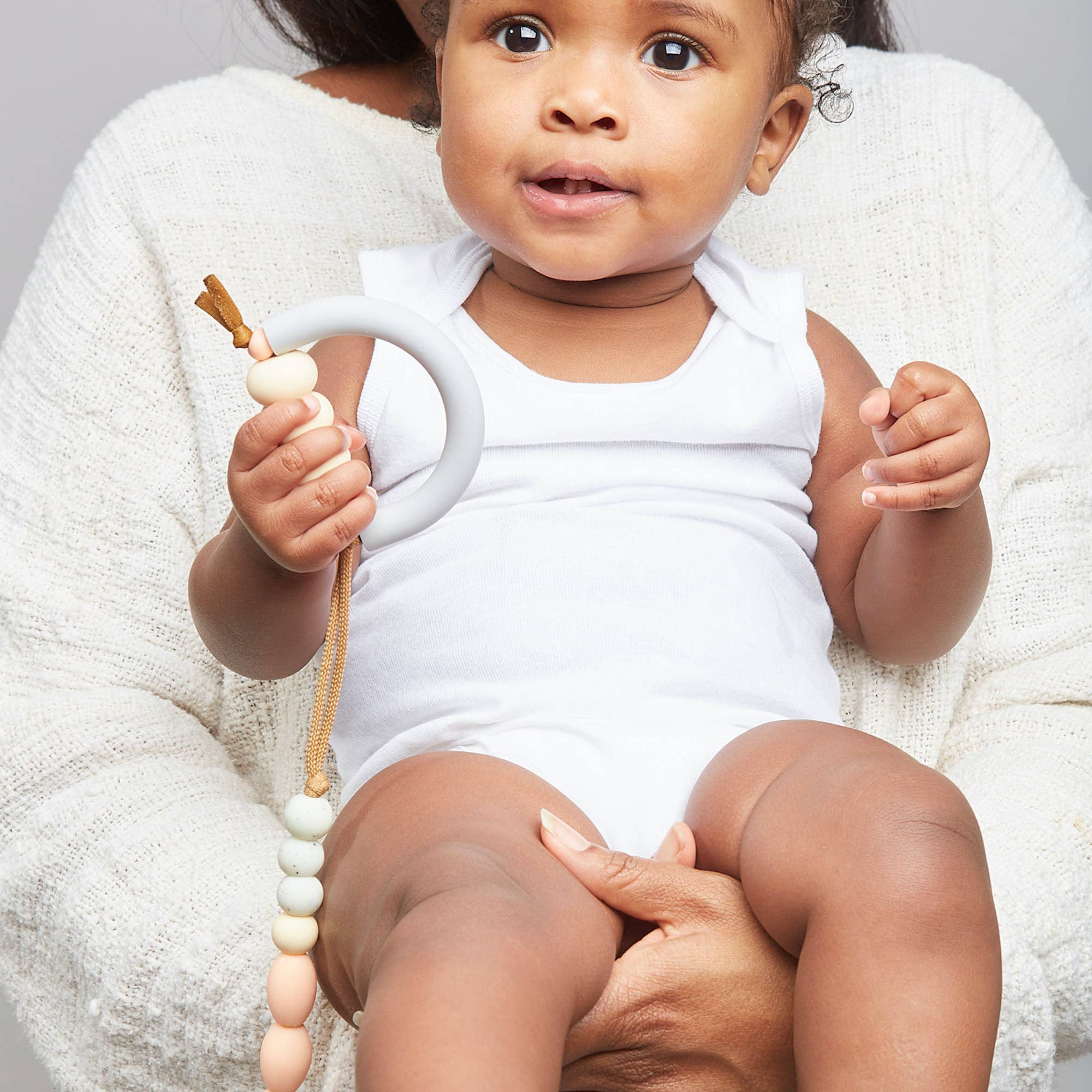 Baby in a white outfit holding a teething ring with a neutral background